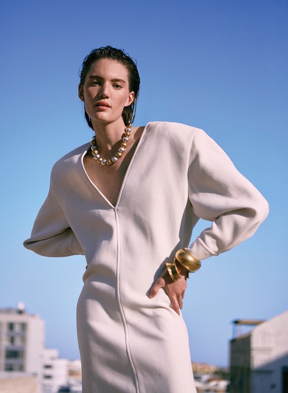 Elegant female model in a cream dress and pearl necklace posing outdoors under a bright blue sky with buildings in the background.
