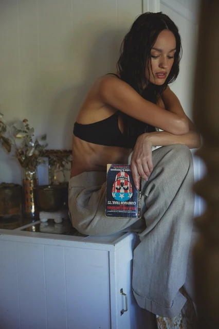 Brunette woman in a black bra and beige pants sits on a white cabinet holding a book with a red, white, and blue cover in an indoor setting.