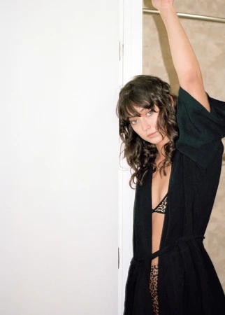 Young woman in a robe with animal-print bra and bottoms stands indoors near a white wall and doorway in soft, natural light.