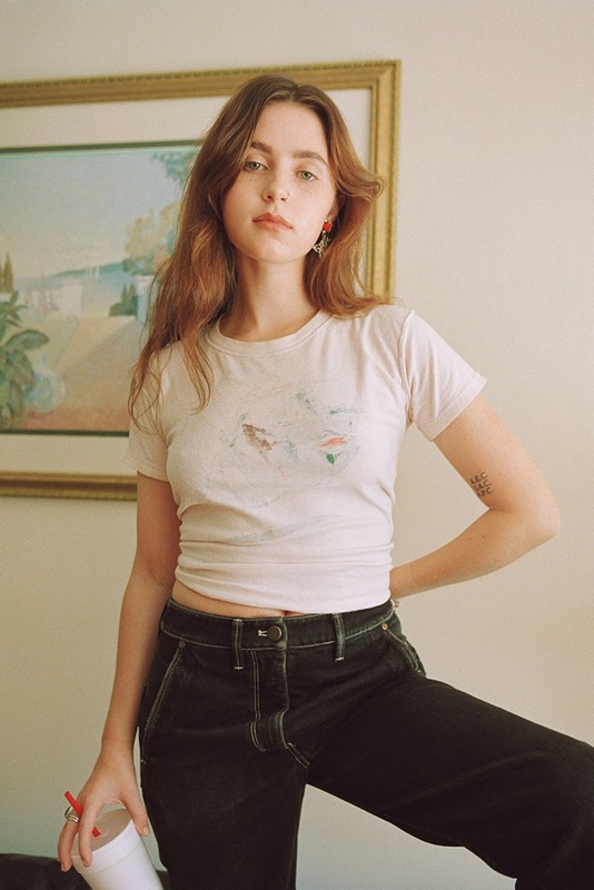 Young woman wearing a white t-shirt and dark jeans posing indoors with a framed picture in the background.