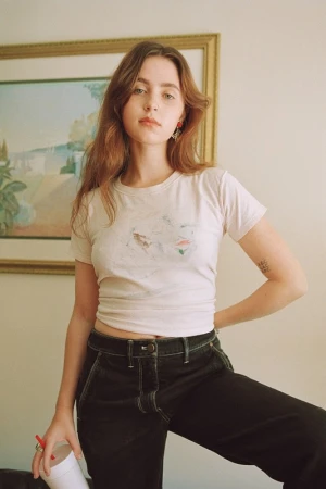 Young woman wearing a white t-shirt and dark jeans posing indoors with a framed picture in the background.