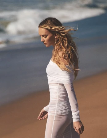 Woman with long blonde hair wearing a sheer white off-the-shoulder dress walking along a sandy beach near ocean waves.