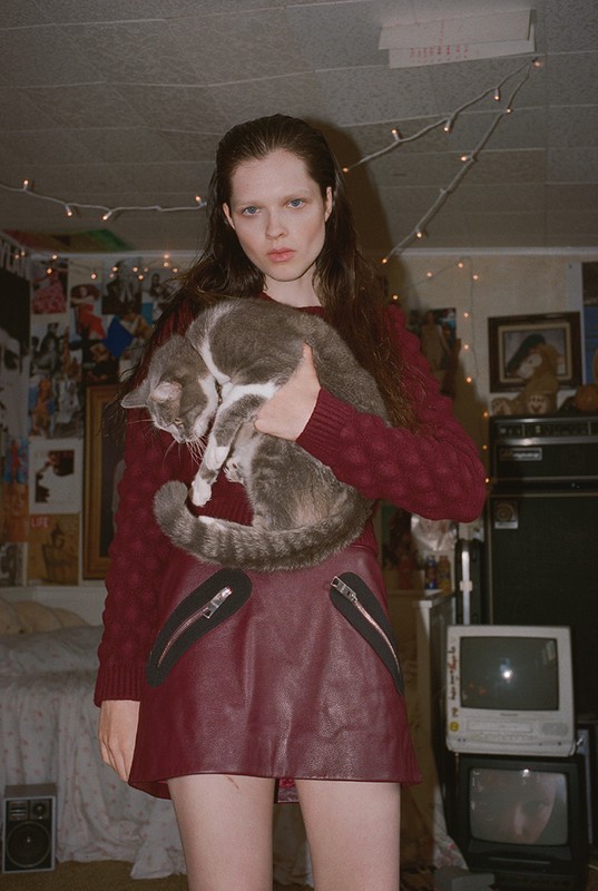 Indoor portrait of a fair-skinned adult female holding a grey and white tabby cat wearing a red sweater and skirt with zippers.