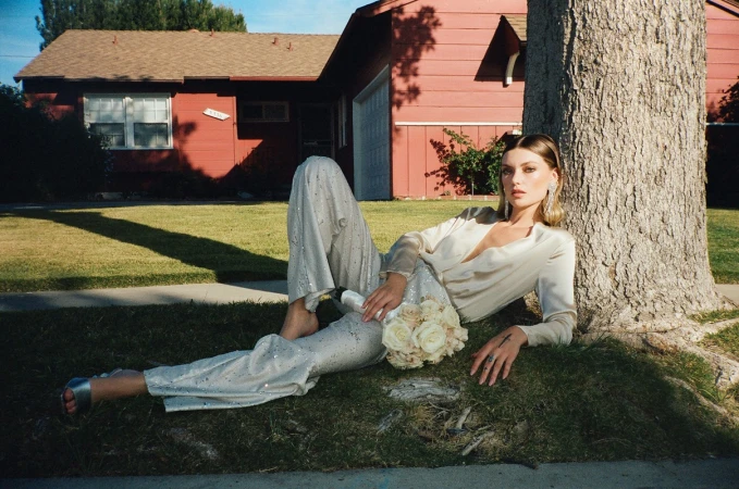 Woman in cream silk pantsuit reclining on grass holding a rose bouquet beside a tree in front of a red house