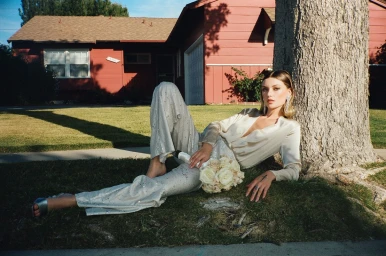 Woman in cream silk pantsuit reclining on grass holding a rose bouquet beside a tree in front of a red house