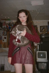 A young woman with long dark hair wearing a maroon textured sweater and leather skirt holds a grey and white cat in a bedroom.
