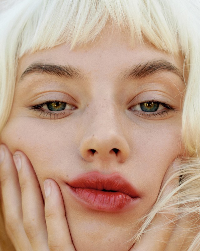Close-up portrait of a white woman with platinum blonde hair framing her face and hands touching her cheeks.