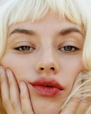 Close-up portrait of a white woman with platinum blonde hair framing her face and hands touching her cheeks.