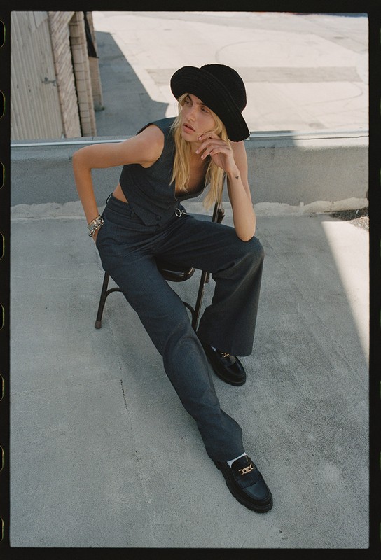 A blonde woman poses outdoors in a black hat and matching vest and pants, seated on a chair with legs outstretched, against a building backdrop.