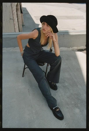 A blonde woman poses outdoors in a black hat and matching vest and pants, seated on a chair with legs outstretched, against a building backdrop.