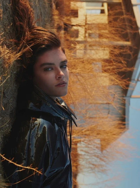 Portrait of a dark-haired woman in a shiny black jacket, lying in golden grass with a building in the background on a sunny day.