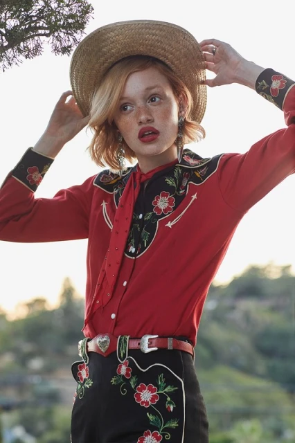 A person wearing a cowboy-inspired outfit and straw hat poses outdoors with greenery visible in the background, shot in natural light.