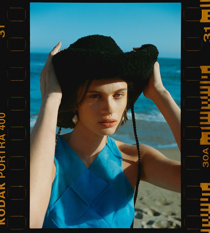 Young woman in blue dress and fuzzy hat posing at the beach on a sunny day; film border visible on sides of image.