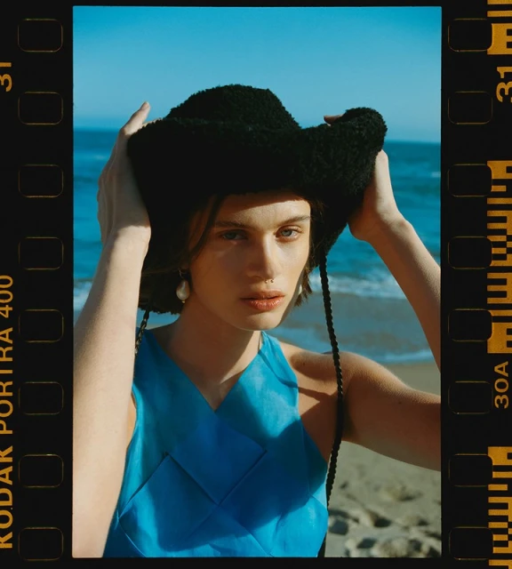 Young woman in blue dress and fuzzy hat posing at the beach on a sunny day; film border visible on sides of image.