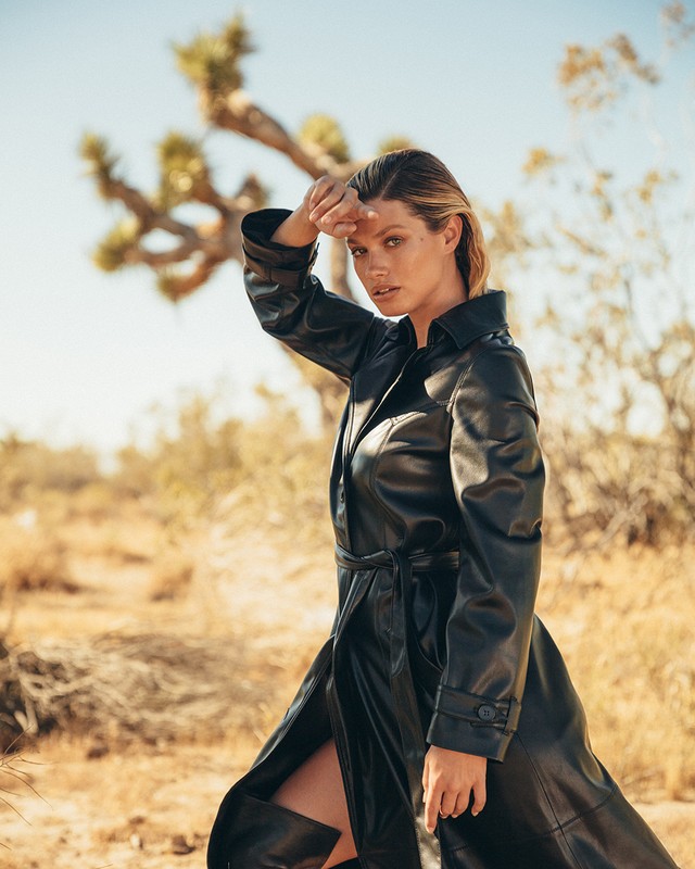 A fashionable woman in a black leather trench coat poses in a desert landscape with a Joshua tree under a clear blue sky.