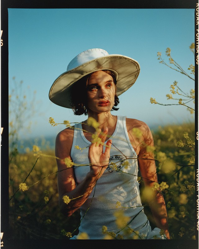 Young woman in white tank top and hat posing in field of yellow wildflowers under bright blue sky, captured in medium outdoor shot.