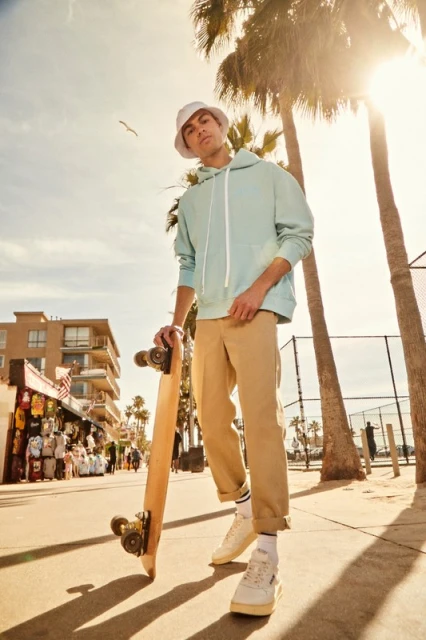 Man in a hoodie and bucket hat holds a skateboard on a sunny street lined with palm trees and shops, showcasing urban style.