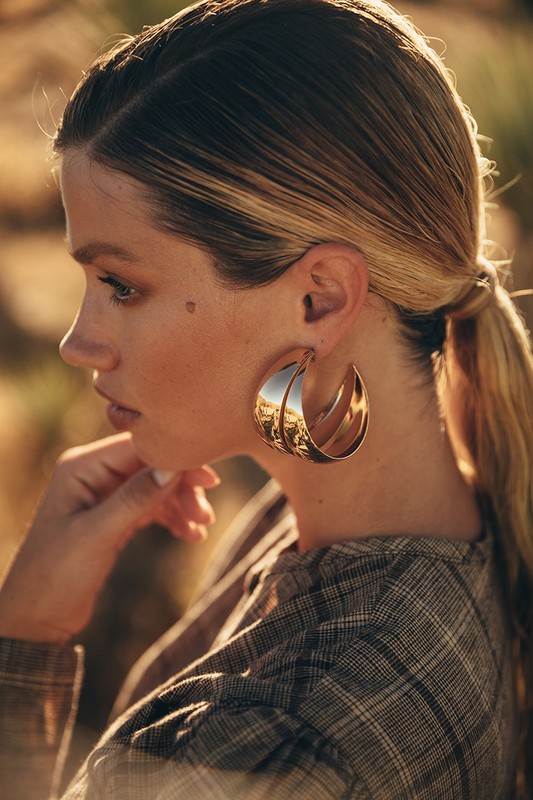 Close-up of a fair-skinned woman with blonde hair in a ponytail wearing large gold earrings and a plaid shirt, with a blurred background.