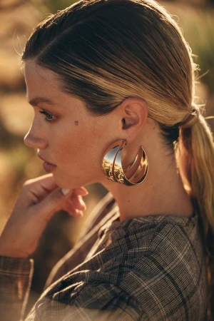 Close-up portrait of a woman wearing large gold hoop earrings and a plaid top with her hair tied in a low ponytail outdoors.