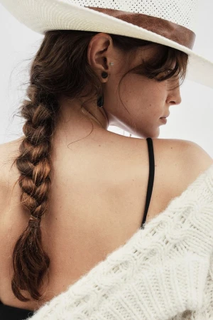 Close-up portrait of a woman wearing a white perforated cowboy hat and a heavy cable-knit sweater, featuring a long braided hairstyle.