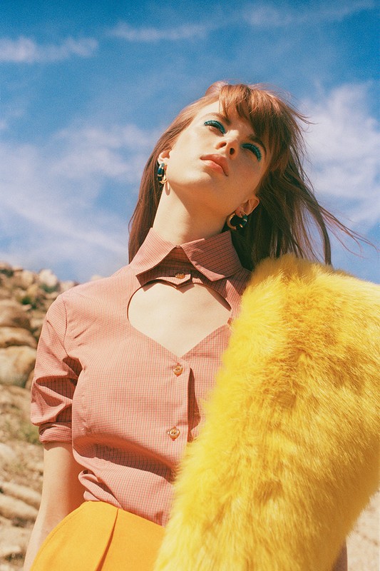 Woman in a red patterned collared shirt with a heart cut-out, standing outdoors in a desert with a blue sky background.