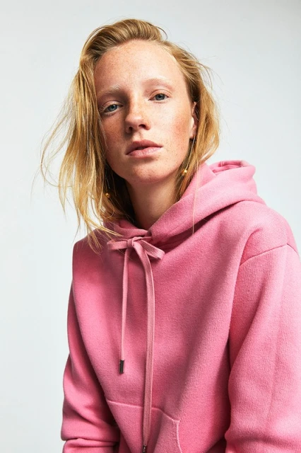 Portrait of a young adult woman with fair skin and freckles wearing a pink hooded sweatshirt against a bright white background in studio lighting.