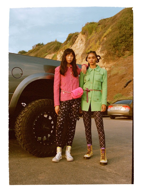 Two female models pose in front of a dark gray truck on a sunny day; one in pink, one in green, with a mountain backdrop.