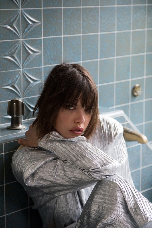 Indoor portrait of a woman with dark hair, wearing a silver shirt and sitting on a tile floor in front of a blue tiled wall.