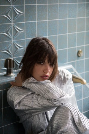 Indoor portrait of a woman with dark hair, wearing a silver shirt and sitting on a tile floor in front of a blue tiled wall.