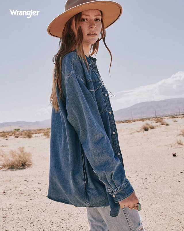 A woman in a denim shirt and hat poses in a desert landscape under a bright sky. The Wrangler logo is visible in the top left.