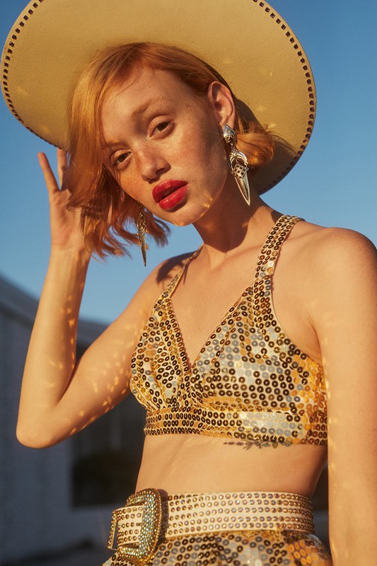A young adult fashion model in a sequined top and cowboy hat, with statement earrings, poses outdoors against a bright blue sky.