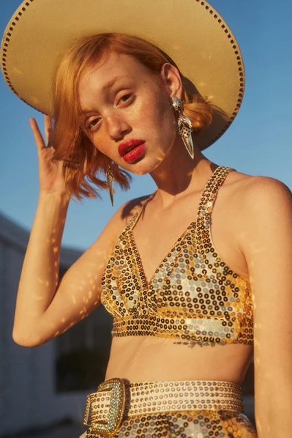 A young adult fashion model in a sequined top and cowboy hat, with statement earrings, poses outdoors against a bright blue sky.