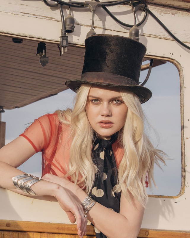 A blonde woman in a top hat and sheer top poses on a boat with sunlight, showcasing vintage fashion and nautical style.