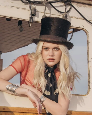 A blonde woman in a top hat and sheer top poses on a boat with sunlight, showcasing vintage fashion and nautical style.