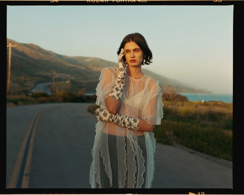 A woman in a sheer white dress and patterned gloves stands on a road with mountains and ocean in the background during golden hour.