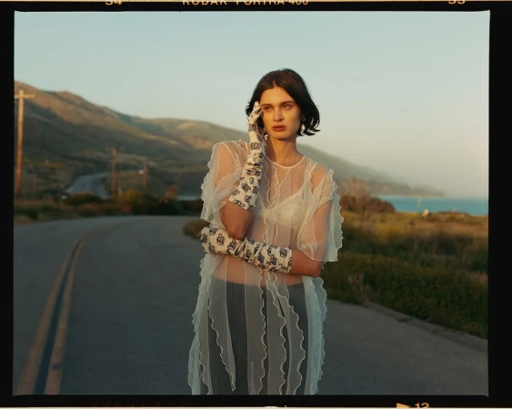 A woman in a sheer white dress and patterned gloves stands on a road with mountains and ocean in the background during golden hour.