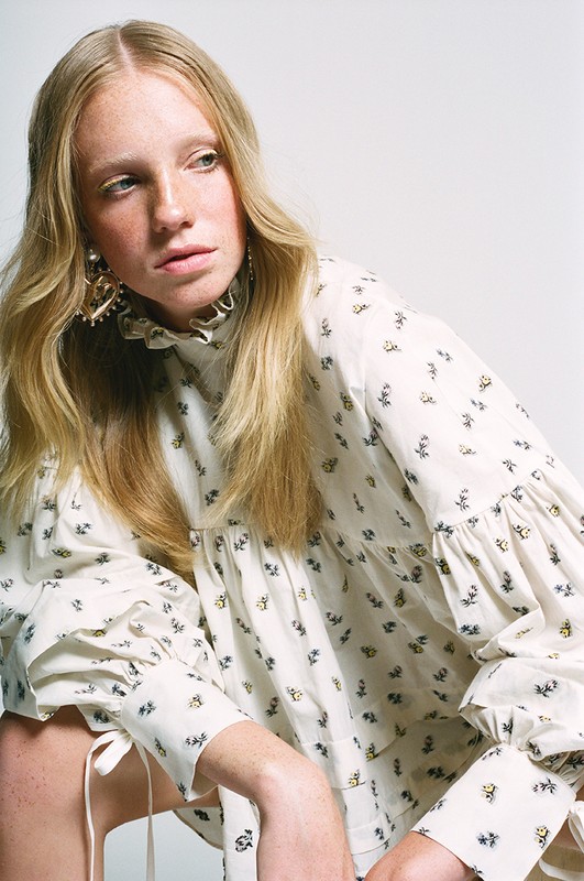 Portrait of fair-skinned woman with freckles wearing a floral blouse and ornate earrings against a white backdrop in a photo studio.