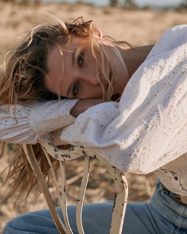 Blonde woman with blue eyes rests on chair in a desert landscape; she wears a white top and jeans in a fashion-style portrait.
