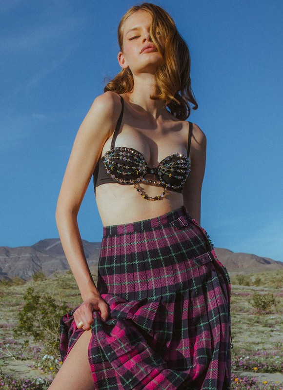 A blonde woman poses in a black bra and plaid skirt in a sunny, arid landscape with mountains and blue sky, showcasing fashion.