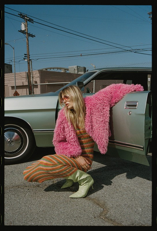 A woman in a pink coat and sunglasses poses next to a silver car in an urban setting, wearing lime green boots and a striped dress.