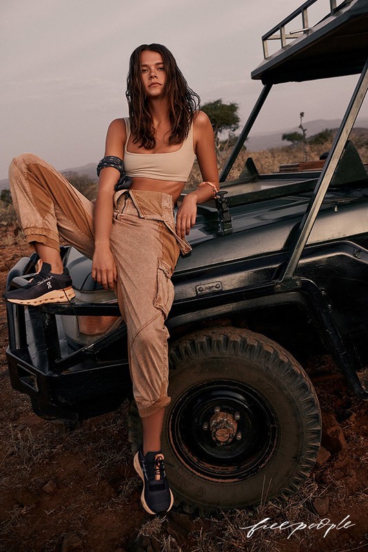 Brunette woman in sports bra and tan pants poses on a black off-road vehicle in a rural setting with dry grassland.