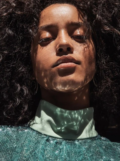 Close-up portrait of a woman with curly hair and green top. Soft dappled sunlight creates shadows on her face and neck.