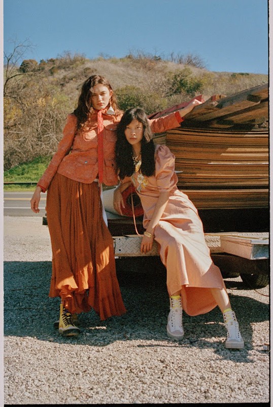 Two women in vintage-style fashion pose outdoors with a pile of lumber under a bright sky, exuding a casual and stylish vibe.