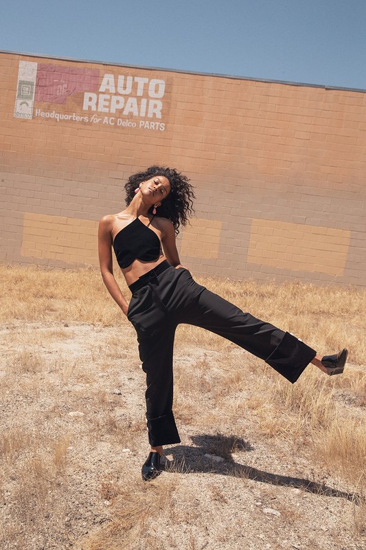 Woman in black halter top and trousers poses in arid landscape near building with faded 'Auto Repair' sign on sunny day.