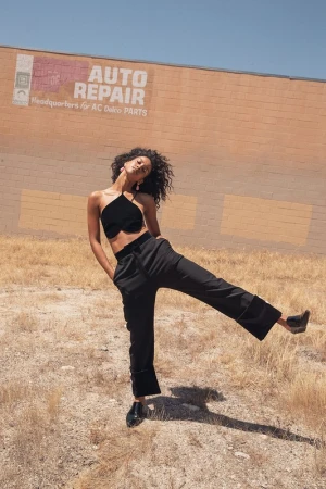 Woman in black halter top and trousers poses in arid landscape near building with faded 'Auto Repair' sign on sunny day.
