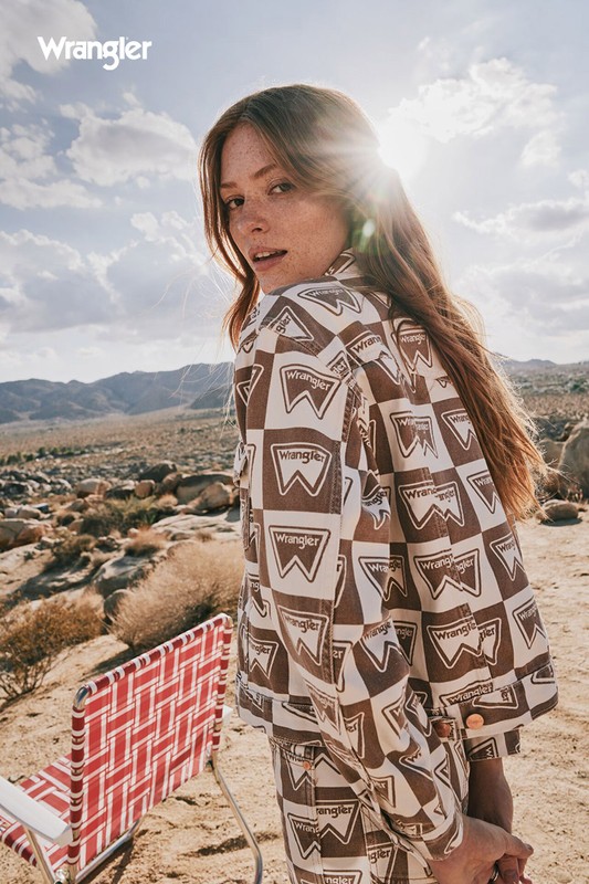 A woman in a patterned jacket stands in a desert landscape with a distant mountain range on a sunny day, wearing Wrangler clothing.