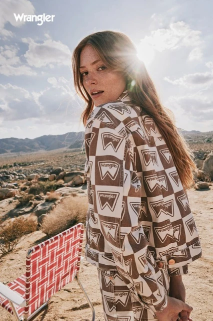 A woman in a patterned jacket stands in a desert landscape with a distant mountain range on a sunny day, wearing Wrangler clothing.