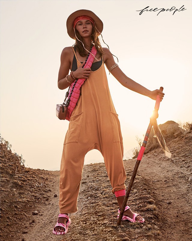 Woman in yellow jumpsuit, sunhat, and sandals hikes on a dirt trail at golden hour, carrying a pink bag and walking stick.