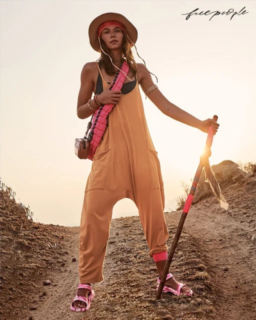Woman in yellow jumpsuit, sunhat, and sandals hikes on a dirt trail at golden hour, carrying a pink bag and walking stick.