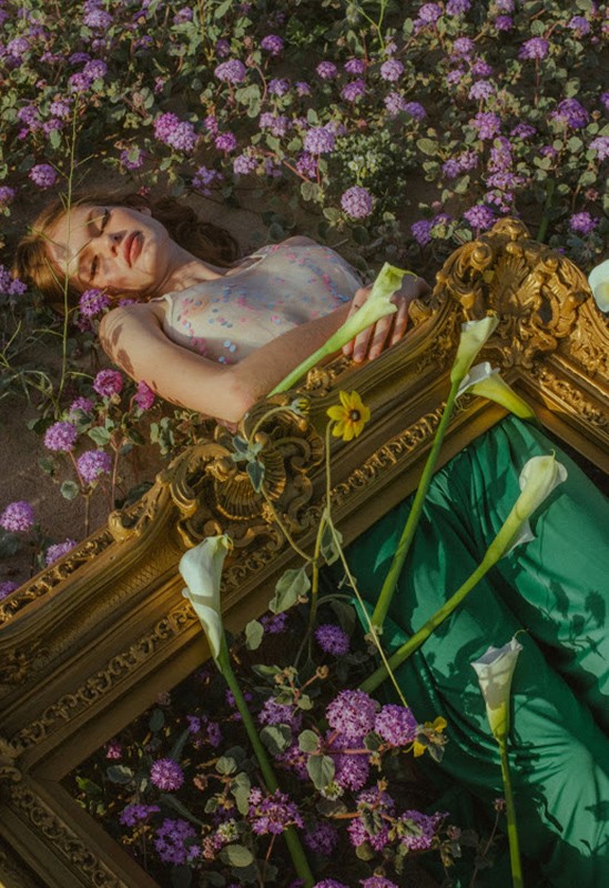 Woman laying on an ornate gold frame in a field of small purple wildflowers. She is surrounded by white calla lily flowers.